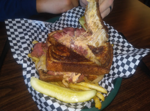 A grilled sandwich with melted cheese and meat, served with fries and a pickle on a checkered paper-lined plate.