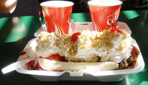 A dessert in a takeout container with banana, whipped cream, nuts, cherries, and two cups of soda in the background.