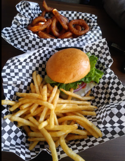 A plate with a burger topped with lettuce and tomato, served with fries and a side of onion rings in checkered paper.