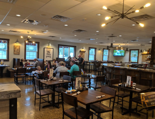 A cozy restaurant interior with wooden tables, patrons dining, and a warm atmosphere. Wall decor includes signs and TVs.