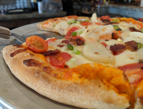 Close-up of a pizza topped with tomatoes, cheese, bacon, and green onions on a rustic wooden table.