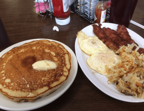 A plate of pancakes with butter, eggs, bacon, and hash browns on a wooden table.