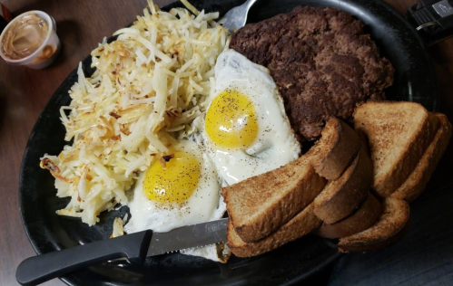 A hearty breakfast plate with two fried eggs, hash browns, a beef patty, and slices of toasted bread.