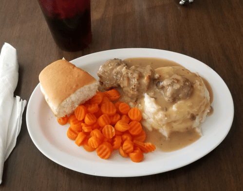 A plate of meatballs in gravy, mashed potatoes, carrots, and a dinner roll, with a drink in the background.