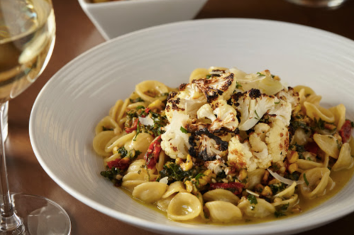 A bowl of orecchiette pasta topped with roasted cauliflower, sun-dried tomatoes, and greens, with a glass of white wine.