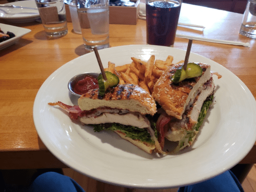 A sandwich with chicken, bacon, lettuce, and pickles, served with fries and dipping sauce on a wooden table.