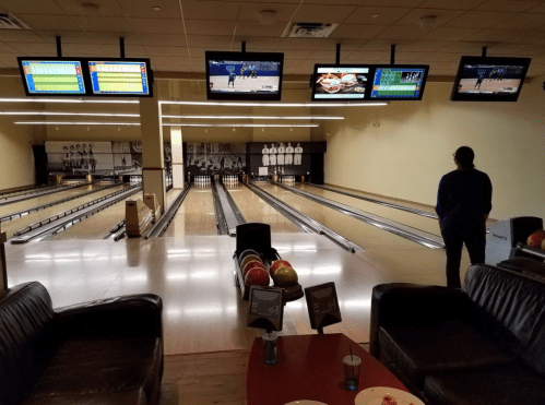 A bowling alley with empty lanes, colorful bowling balls, and multiple screens displaying scores and games.