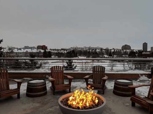 A cozy outdoor seating area with wooden chairs around a fire pit, overlooking a snowy landscape and distant buildings.