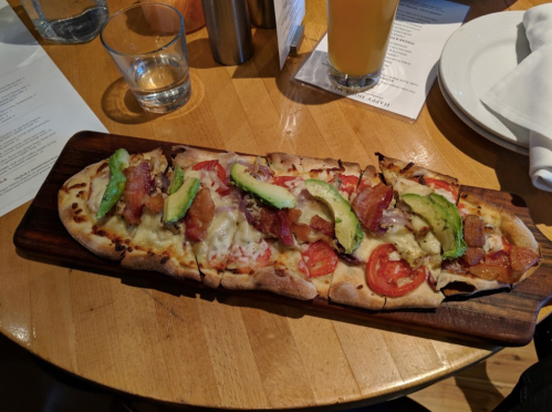 A flatbread pizza topped with cheese, bacon, tomatoes, red onions, and avocado, served on a wooden board.