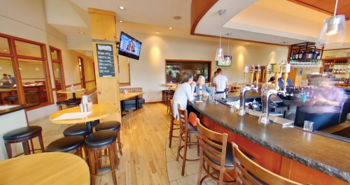 A spacious bar area with wooden tables, stools, and a counter, featuring TVs and a variety of drinks on display.