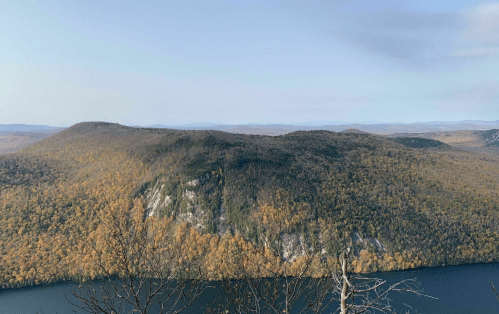 A panoramic view of a mountain covered in autumn foliage, overlooking a calm lake under a clear sky.