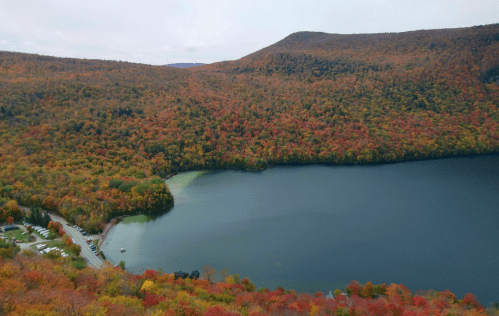Aerial view of a lake surrounded by vibrant autumn foliage and rolling hills under a cloudy sky.
