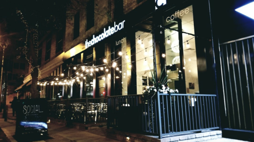 A cozy café at night, featuring outdoor seating and string lights, with a sign reading "thechocolatebar" and "coffee."