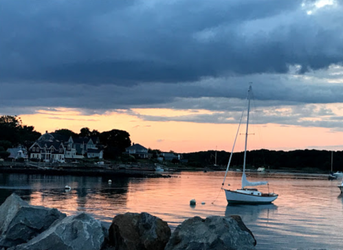 A serene sunset over a calm harbor with a sailboat and charming houses along the shore.