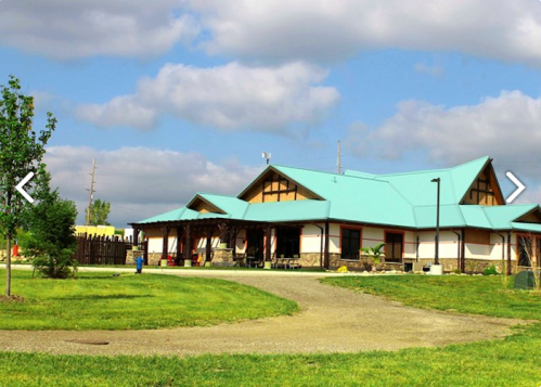 A modern building with a teal roof, surrounded by green grass and trees, under a partly cloudy sky.