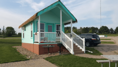 A small turquoise cabin with a thatched roof, white porch, and steps, surrounded by grass and a gravel path.