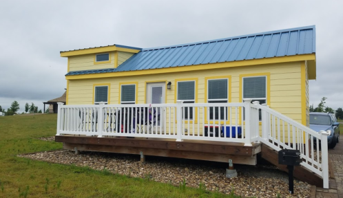 A bright yellow house with a blue metal roof and a white deck, set in a grassy area under a cloudy sky.