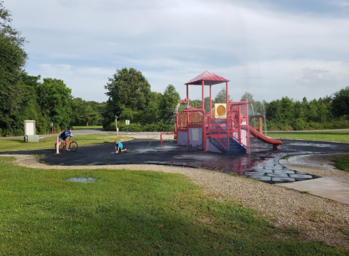 Children playing at a colorful playground with a slide, surrounded by wet ground and green grass under a cloudy sky.