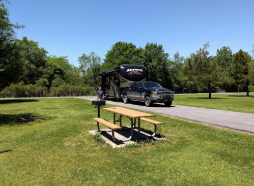 A truck towing an RV parked near a picnic table in a grassy area with trees under a clear blue sky.