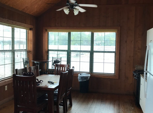 Cozy wooden dining area with a table, chairs, and large windows, featuring a ceiling fan and kitchen appliances.