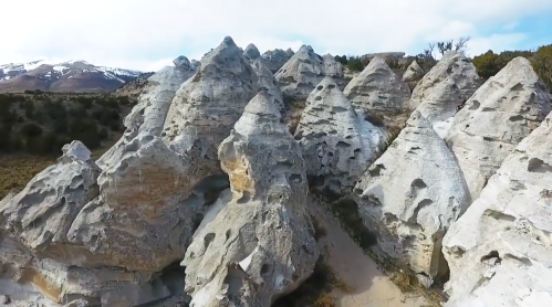 Aerial view of unique, jagged rock formations with a mountainous backdrop and scattered vegetation.