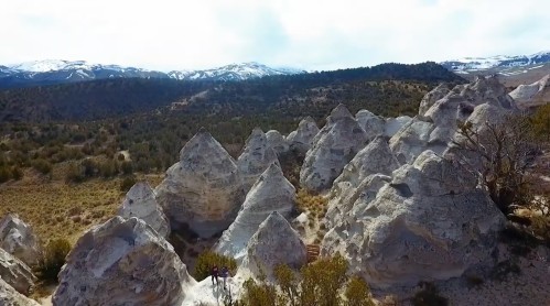 Aerial view of unique rock formations surrounded by greenery and mountains in the background, with snow-capped peaks.