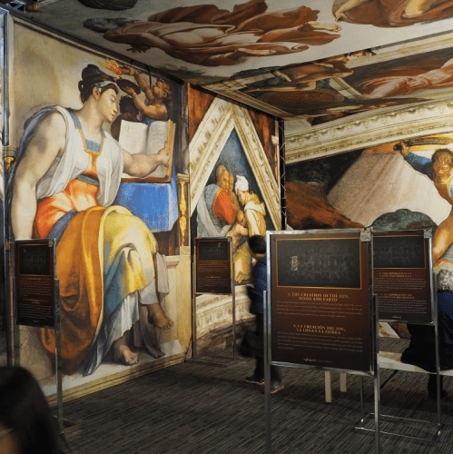 A mural depicting figures from the Creation of Adam, with informational panels in a gallery setting.