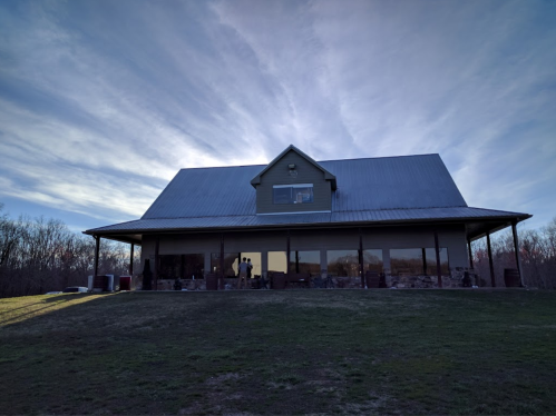 A large green house with a metal roof, set against a cloudy sky and surrounded by trees.