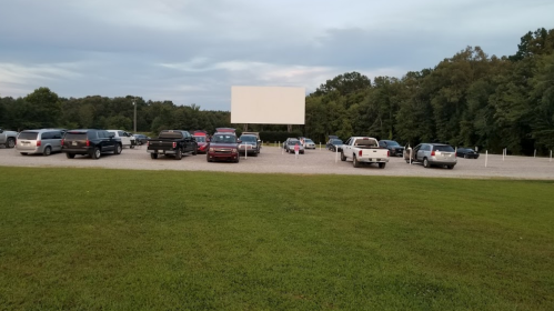 A drive-in movie theater with parked cars and a blank screen, surrounded by trees under a cloudy sky.