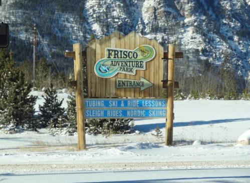Sign for Frisco Adventure Park, listing activities: tubing, ski lessons, sleigh rides, and Nordic skiing. Snowy landscape.