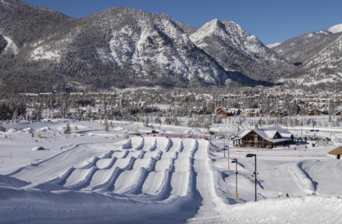 Snow-covered landscape with ski slopes and a lodge, surrounded by mountains under a clear blue sky.
