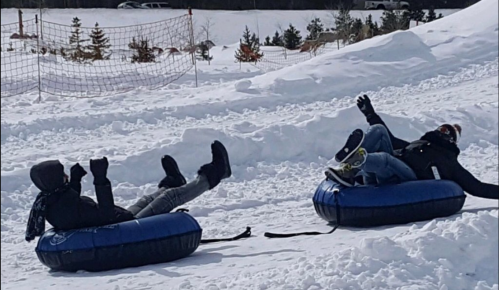 Two people in winter clothing are sitting on blue snow tubes, laughing and enjoying a snowy hill.