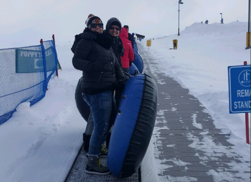 Two people stand on a snowy slope, holding inflatable tubes, ready to go tubing. Snowy landscape in the background.