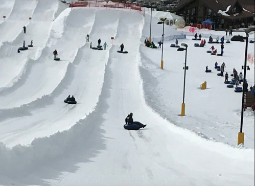 A snowy tubing hill with people sliding down on inflatable tubes, surrounded by snow-covered slopes and a lodge in the background.