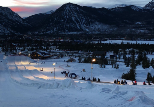 A snowy landscape at dusk, featuring a sledding hill with people enjoying the winter scenery and mountains in the background.