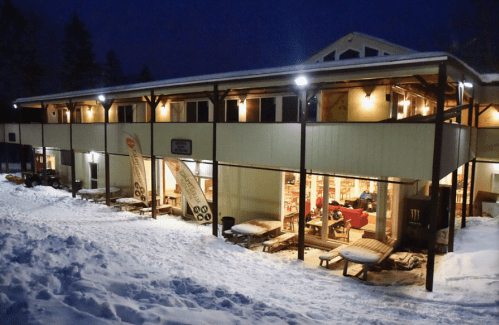 A cozy building at night, illuminated with lights, surrounded by snow and featuring outdoor seating.