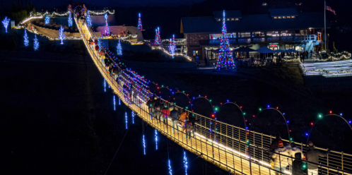 A brightly lit suspension bridge at night, adorned with colorful holiday lights, with people walking across it.