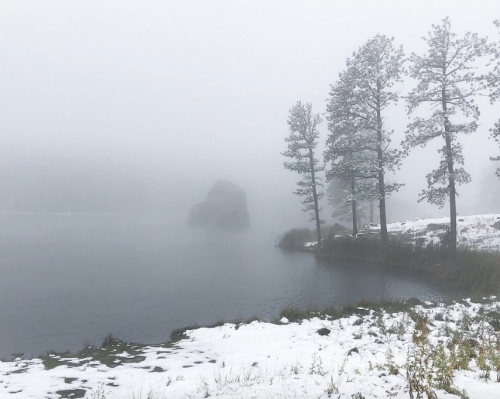 A foggy landscape featuring a lake, a large rock, and pine trees, with snow on the ground.
