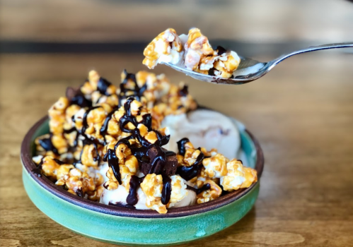 A bowl of ice cream topped with caramel popcorn and drizzled with chocolate sauce, with a spoon lifting some popcorn.