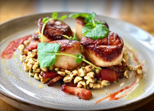 Seared scallops on a bed of corn and bacon, garnished with microgreens, served on a decorative plate.