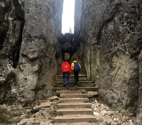 Two people walk up stone steps between tall, rugged rock formations in a natural setting.