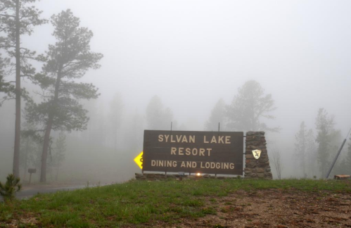 A foggy scene featuring a wooden sign for Sylvan Lake Resort, indicating dining and lodging, surrounded by trees.