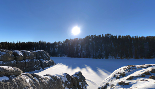 A snowy landscape with a frozen lake, rocky foreground, and a bright sun shining over a forested hillside.