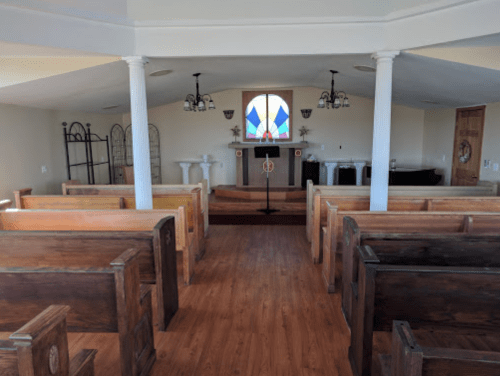 Interior of a small chapel with wooden pews, a podium, and a stained glass window at the front.