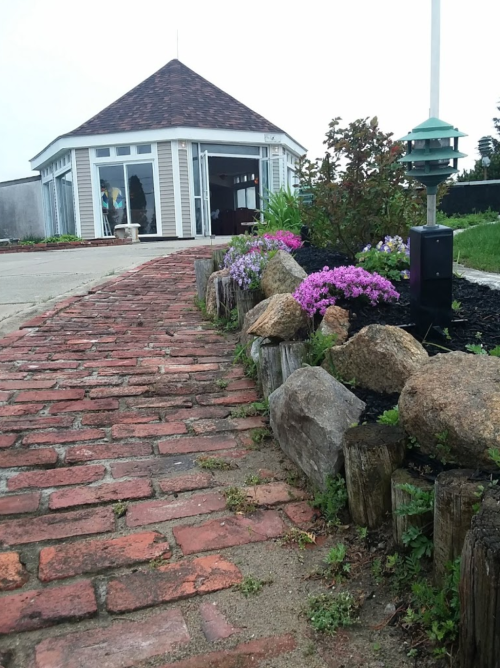 A brick pathway lined with flowers and rocks leads to a round building with a conical roof.