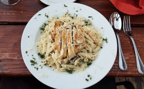 A plate of penne pasta topped with grilled chicken and sprinkled with grated cheese, garnished with parsley.