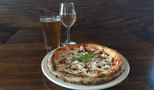 A pizza topped with mushrooms and onions, served with a glass of beer and a glass of white wine on a wooden table.