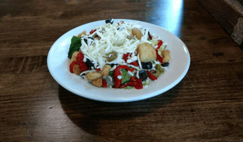 A colorful salad with mixed greens, black olives, red peppers, croutons, and shredded cheese on a white plate.