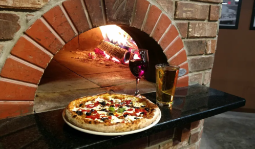 A freshly baked pizza with toppings, a glass of red wine, and a beer, set against a brick pizza oven.