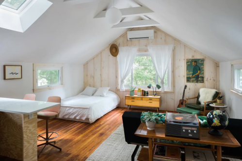 Cozy attic room with a bed, seating area, and natural light, featuring wooden walls and stylish decor.
